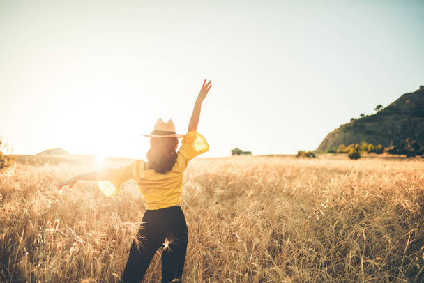 Chica joven vestida de amarillo con sombrero de paja se encuentra feliz en el medio del campo de trigo al atardecer. - Foto, Imagen