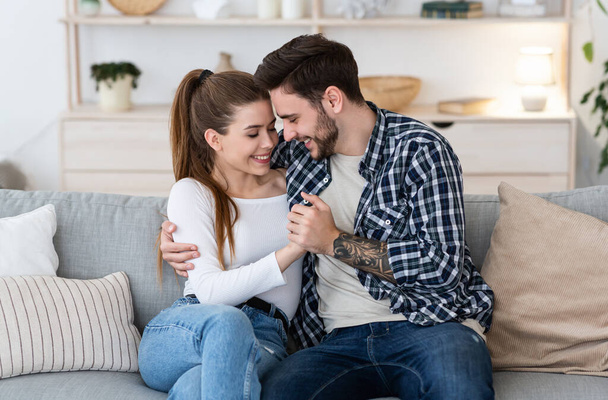 Felices juntos en casa. Feliz joven hombre y mujer en ropa casual están tomados de la mano, abrazándose en el sofá - Foto, imagen