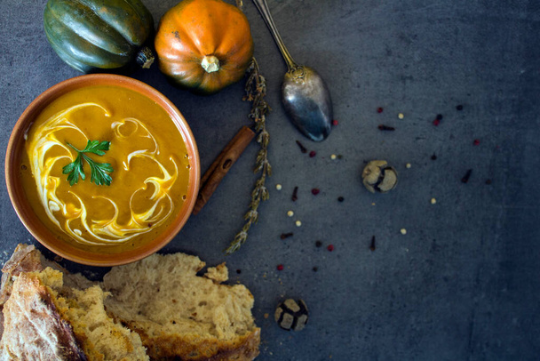 灰色のテーブルの上に焼きたてのパンと銀のスプーンでカボチャのスープ.ビーガンメニューのトップビューの写真。健康的な食事の概念。暗いテクスチャの背景とコピースペース.  - 写真・画像