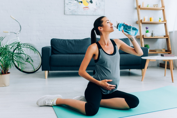 Mujer embarazada bebiendo agua mientras entrena en la esterilla de fitness en casa  - Foto, Imagen