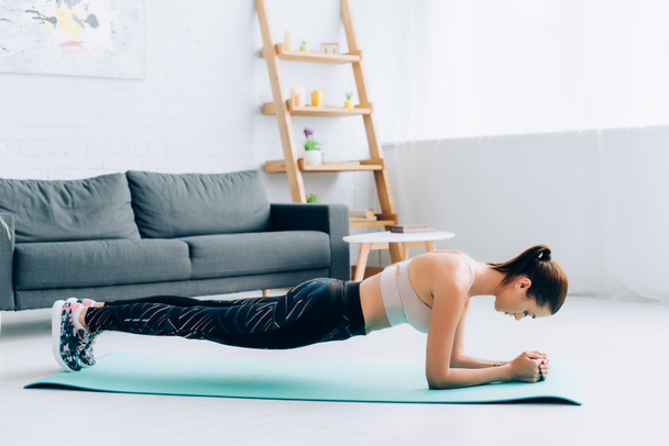 Side view of sportswoman standing in plank on fitness mat in living room  - Photo, Image