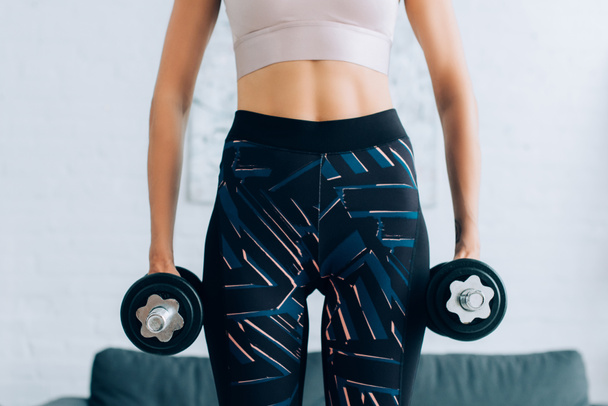Cropped view of fit sportswoman holding dumbbells at home  - Foto, afbeelding