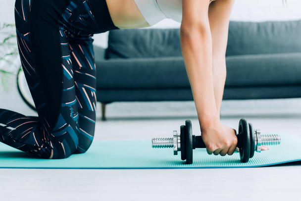 Cropped view of sportswoman holding dumbbell on fitness mat at home  - Fotografie, Obrázek