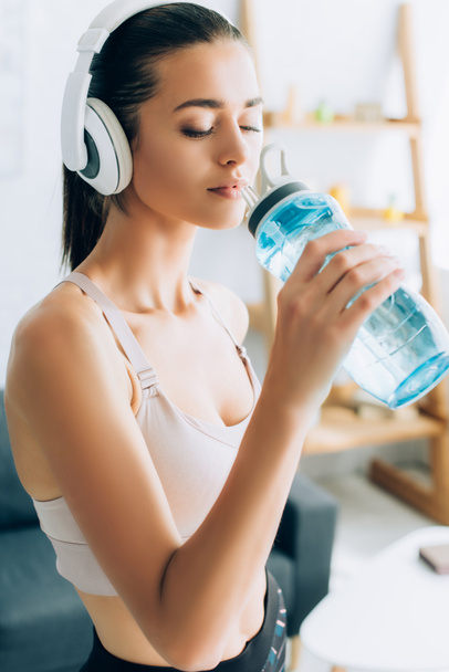 Mujer joven con auriculares sosteniendo botella deportiva mientras entrena en casa  - Foto, imagen