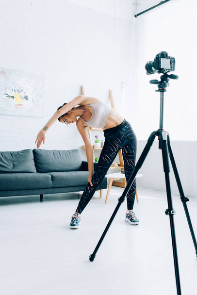 Selective focus of young sportswoman stretching near digital camera in living room  - Photo, Image
