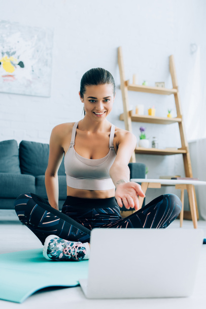 Selective focus of young sportswoman pointing with hand at laptop while sitting on fitness mat  - Photo, Image