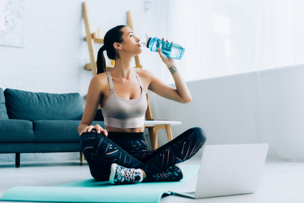 Selektiver Fokus der Sportlerin, die Wasser trinkt, während sie in der Nähe des Laptops auf der Fitnessmatte sitzt  - Foto, Bild