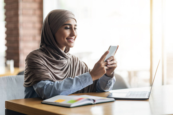 Jeune femme en hijab avec ordinateur portable en utilisant un smartphone au café - Photo, image