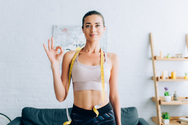 Brunette sportswoman with measuring tape around neck showing okay gesture at home  - Photo, Image