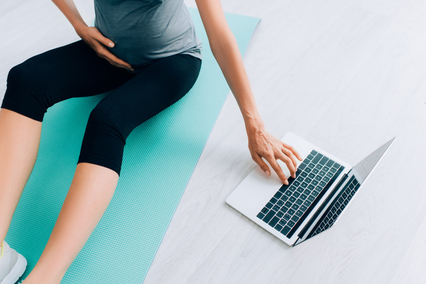 Cropped view of pregnant woman using laptop on fitness mat at home  - Фото, изображение