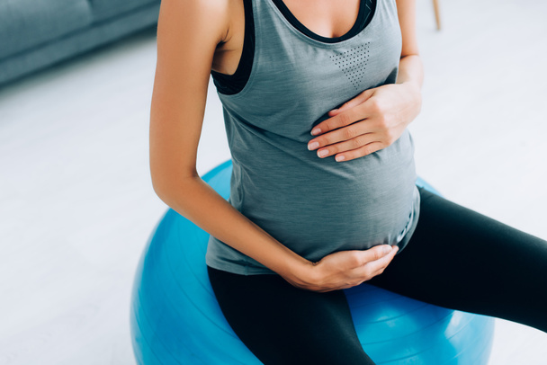 Cropped view of pregnant woman in sportswear touching belly on fitness ball at home  - Photo, Image