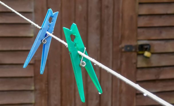 Blue plastic clothes peg hangs upside down on a washing line after a rain shower. Similar green one deliberately out of focus in foreground. Water droplets, with rustic wooden shed background. England - Photo, Image