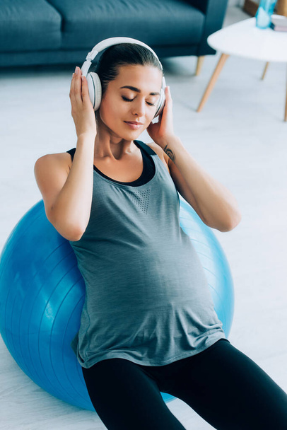 Young pregnant woman listening music in headphones while exercising with fitness ball at home  - Fotó, kép