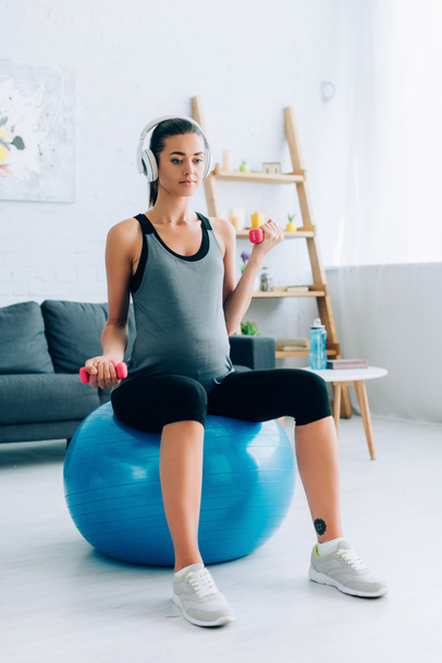 Zwangere sportvrouw in hoofdtelefoon uit te werken met halters en fitness bal thuis  - Foto, afbeelding