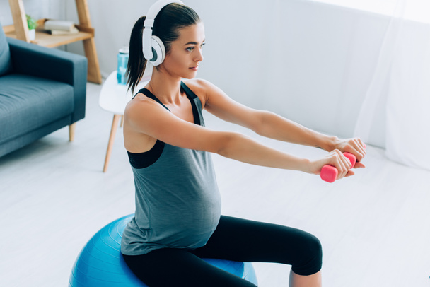 Pregnant sportswoman training with dumbbells and fitness ball while listening music in headphones  - Photo, Image
