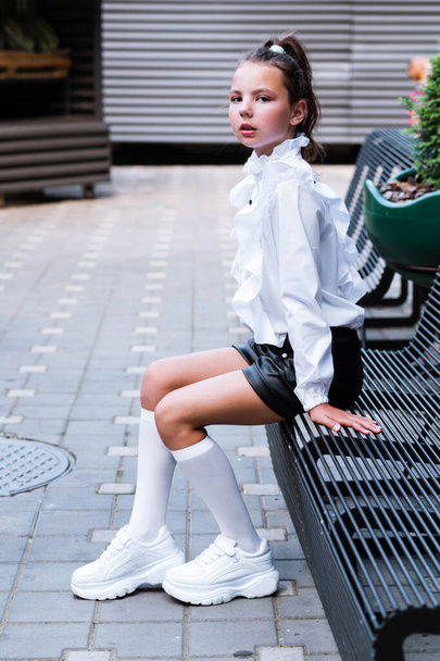 Una colegiala hermosa y de moda en una blusa blanca, pantalones cortos negros, calcetines y zapatos deportivos se sienta en un banco de hierro negro. Foto vertical - Foto, Imagen