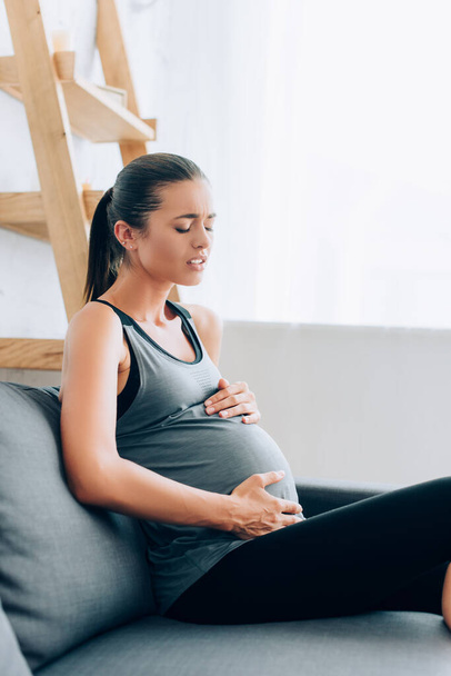 Focus selettivo della giovane sportiva incinta che soffre di dolore sul divano a casa  - Foto, immagini