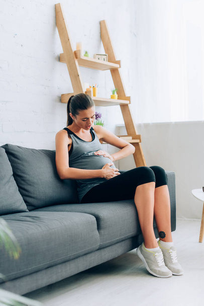 Selective focus of pregnant sportswoman feeling pain and touching belly on couch  - Photo, Image