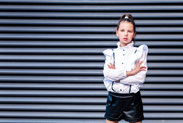 Stylish schoolgirl in a white blouse, black shorts posing against the backdrop of metal blinds. Horizontal photo - 写真・画像