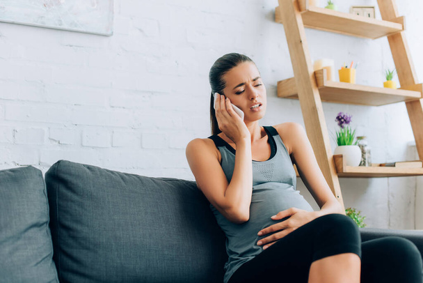 Selective focus of pregnant sportswoman taking on smartphone while feeling pain on couch  - Foto, imagen