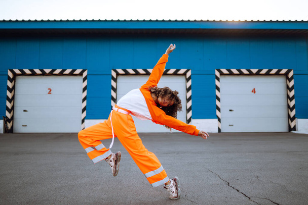 Nuori afroamerikkalainen nainen - tanssija tanssimassa kadulla auringonlaskun aikaan. Tyylikäs nainen, jolla on kiharat hiukset oranssissa puvussa. Urheilu, tanssi ja kaupunkikulttuuri. - Valokuva, kuva