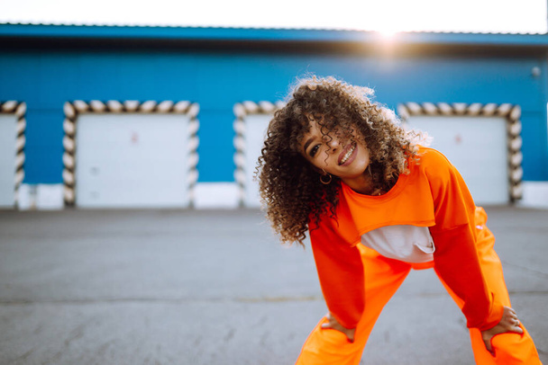 Stylish woman with curly hair in an orange suit posing with a skateboard. African American woman stands on the street with skateboard in hands. Lifestyle, youth concept. - Фото, изображение