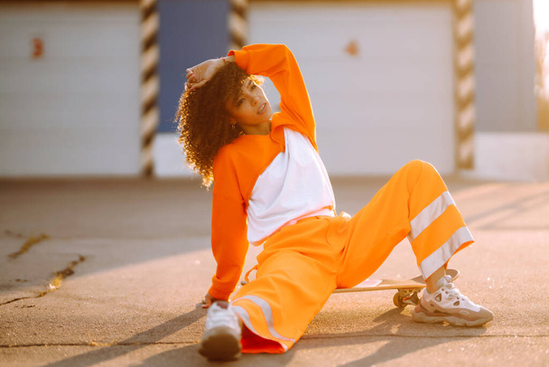 Молодая афроамериканка - танцовщица, танцующая на улице на закате. Стильная женщина с вьющимися волосами в оранжевом костюме, демонстрирующая некоторые движения. Концепция спорта, танцев и городской культуры. - Фото, изображение