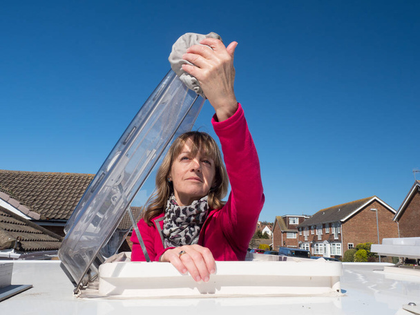 A lady motorhome owner cleans the skylight hatch of her vehicle.The top half of her body is visible as she has climbed through the roof skylight.Vivid pink fleece.Amusing - Photo, Image