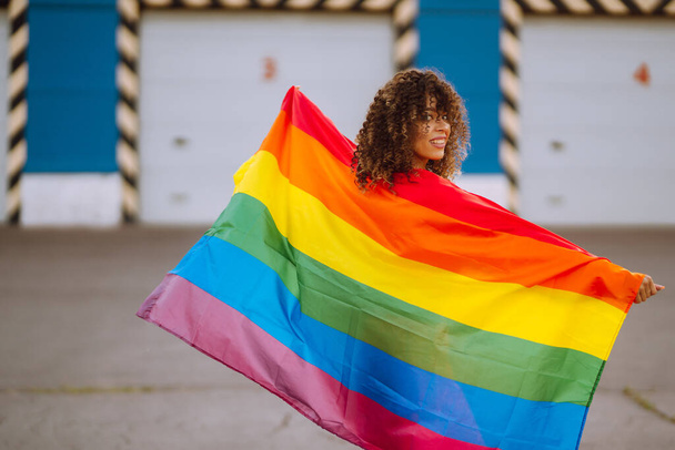 Young African American lesbian woman with LGBT rainbow flag in the street at sunset. Stylish woman with curly hair in an orange suit posing with lgbt pride flag. - Фото, изображение
