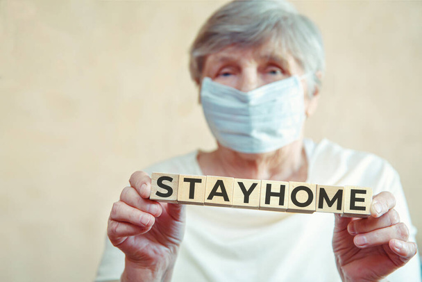 Starší žena s lékařskou maskou na obličeji drží kostky s nápisem STAY HOME. Koncept - karanténa, izolace, karonovirus Covid-19 - Fotografie, Obrázek