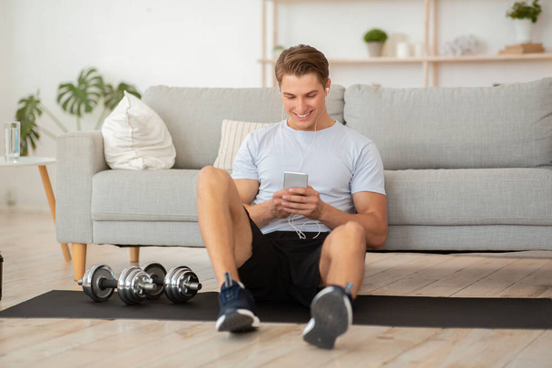 Attractive young man with headphones in sportswear sits on mat on floor with dumbbells and looks at smartphone - Photo, image