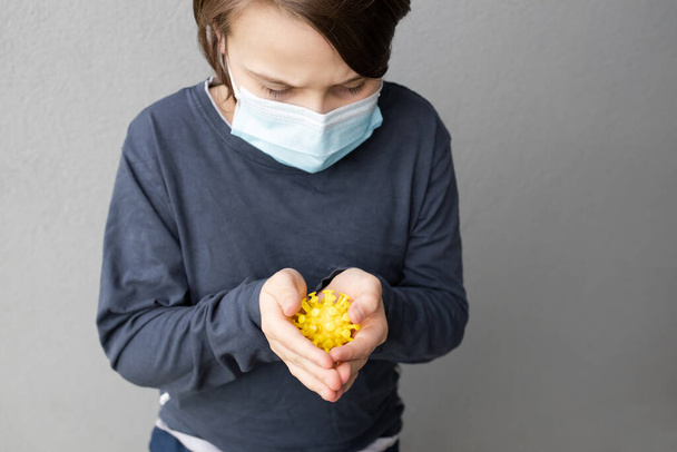 Giovane ragazzo caucasico indossando una maschera chirurgica blu e tenendo un modello di virus corona durante la pandemia COVID-19 - Foto, immagini