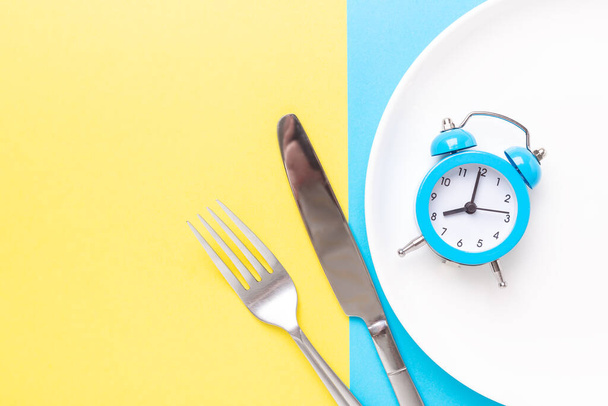Blue alarm clock, fork, knife and empty plate on colored paper background. Intermittent fasting concept - Image - Fotografie, Obrázek