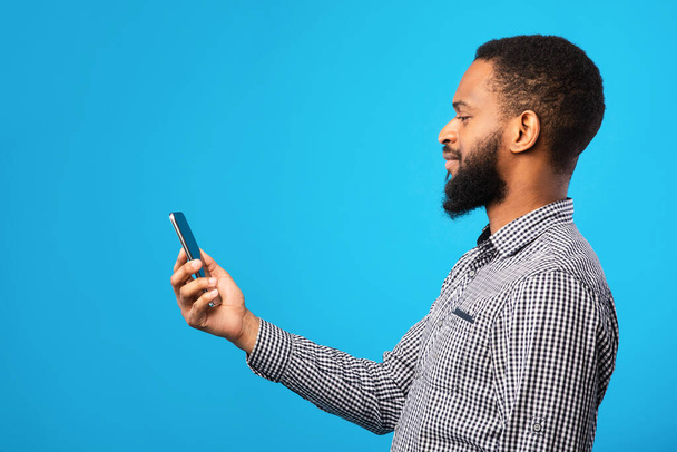 Afro άνθρωπος χρησιμοποιώντας το κινητό τηλέφωνο σε μπλε τοίχο, πλαϊνή άποψη - Φωτογραφία, εικόνα