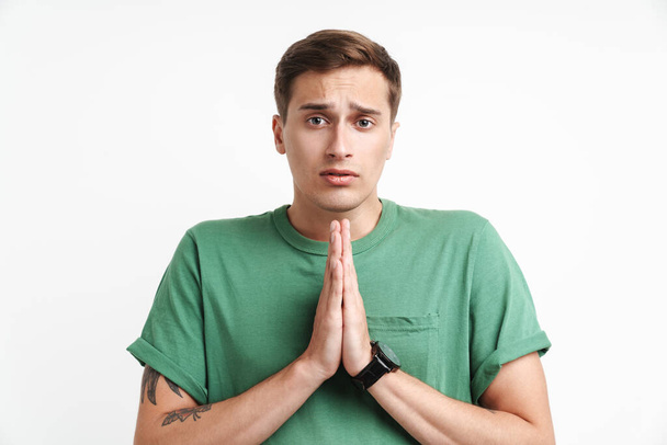 Imagen del hombre caucásico tenso en camiseta básica rezando y sosteniendo las palmas juntas aisladas sobre fondo blanco - Foto, Imagen