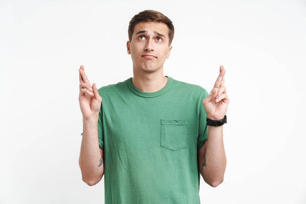 Image of focused caucasian man in basic t-shirt praying and keeping fingers crossed isolated over white background - Foto, Imagen