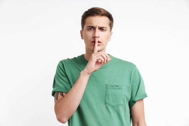 Image of attractive caucasian man in basic t-shirt holding index finger at lips isolated over white background - Foto, Imagem
