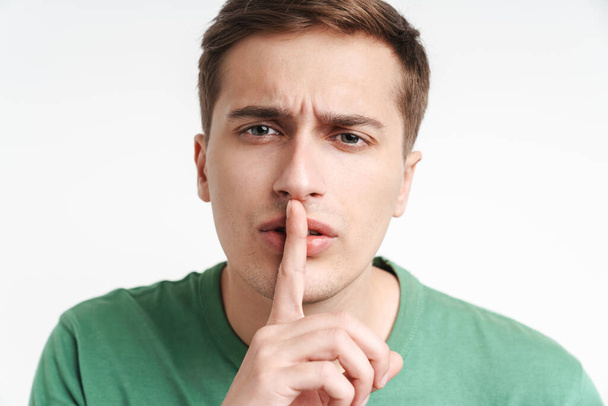 Image of brunette caucasian man in basic t-shirt holding index finger at lips isolated over white background - 写真・画像