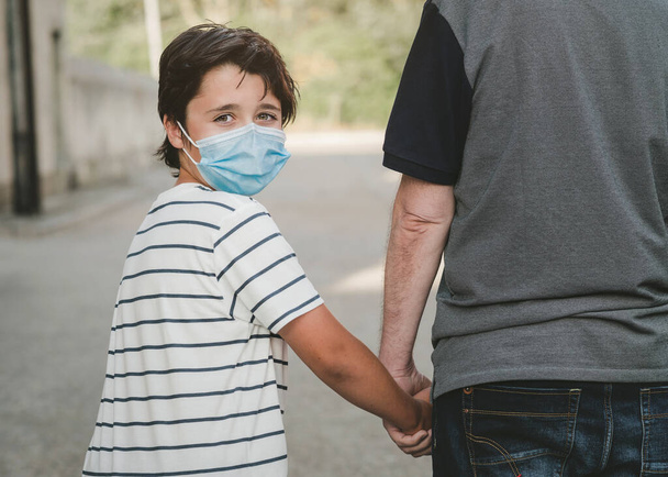 счастливый ребенок в медицинской маске держа за руку своего отца на открытом воздухе - Фото, изображение