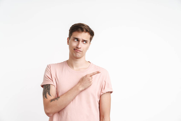Image of displeased young man in basic t-shirt pointing finger aside isolated over white background - Photo, image