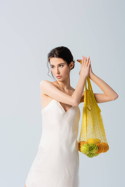 donna bruna in abito di seta tenendo borsa stringa con frutti maturi su bianco, concetto di ecologia  - Foto, immagini