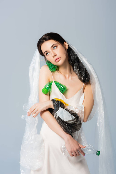 woman with plastic bag on head standing in silk dress with empty bottles isolated on grey, ecology concept   - Photo, Image