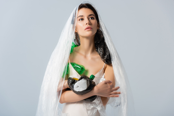 young woman with plastic bag on head standing in silk dress with empty bottles isolated on grey, ecology concept   - Foto, immagini