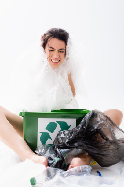 femme pieds nus avec migraine enveloppée dans un sac en plastique assis près de la poubelle avec panneau de recyclage sur blanc, concept d'écologie  - Photo, image