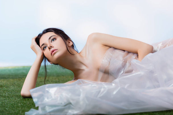 молодая женщина, завернутая в полиэтилен, лежит на зеленой траве и смотрит в сторону синей, экологической концепции  - Фото, изображение