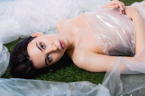 young woman wrapped in polyethylene looking at camera and lying on grass, ecology concept  - Photo, Image