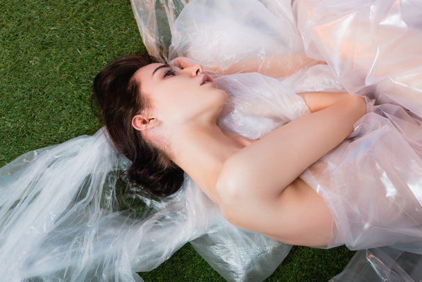 Draufsicht auf junge Frau in Polyethylen gehüllt auf frischem Gras liegend, ökologisches Konzept  - Foto, Bild