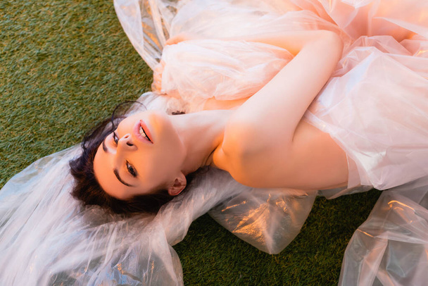 top view of young brunette woman wrapped in polyethylene lying on grass, ecology concept  - Photo, Image