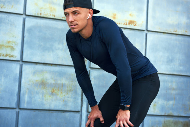 Outdoor shot of athlete man leaning forward with hands on knees and looking away standing against urban metal wall in the city. Sporty male taking a break after running in the city street outside. - Foto, immagini