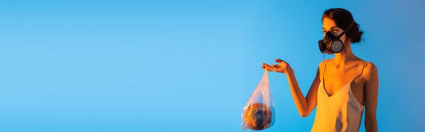 horizontal image of woman in gas mask holding plastic bag with globe on blue - Фото, изображение